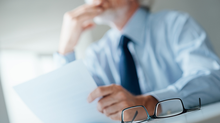 image of manager pondering while holding a piece of paper and eyeglasses set on desk